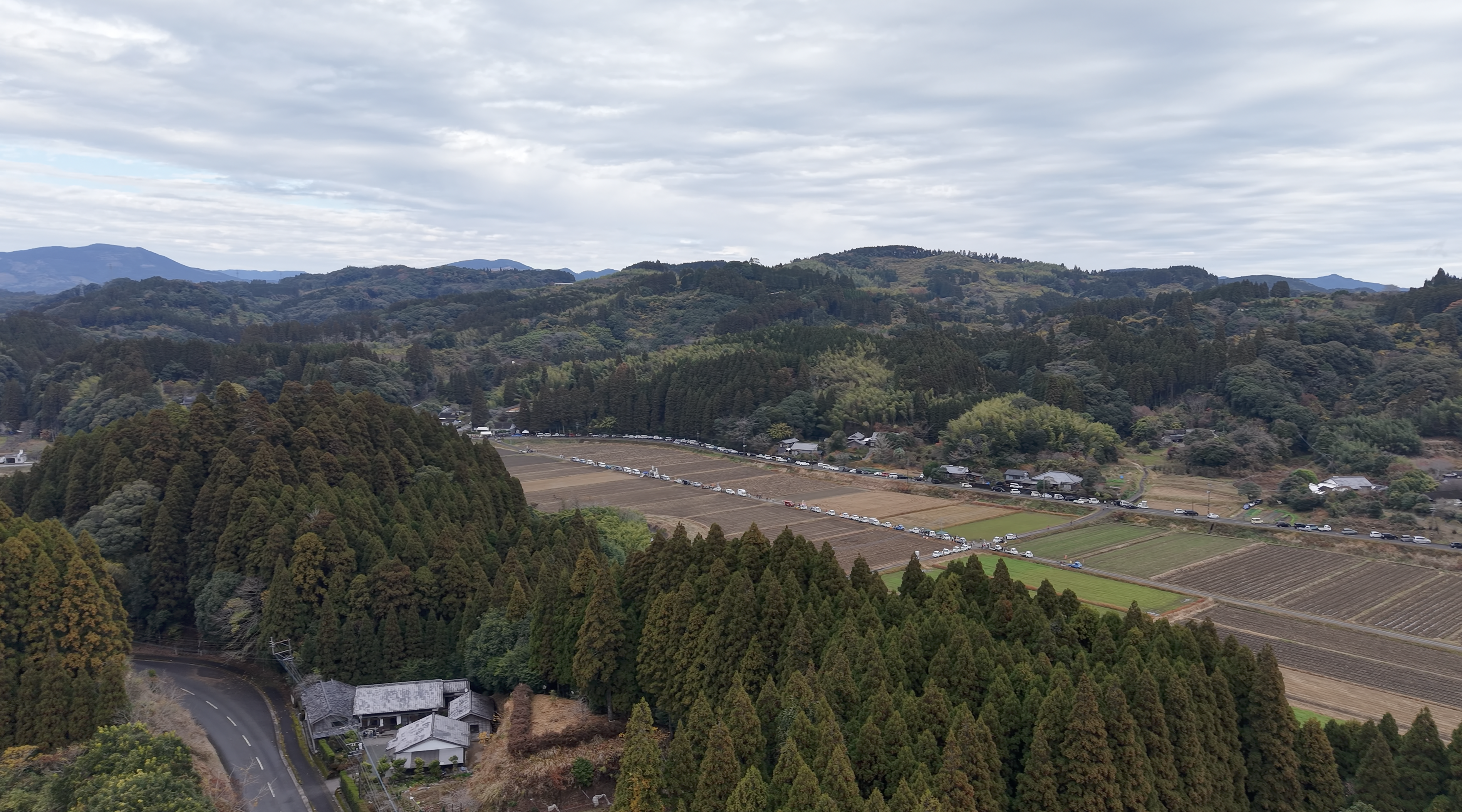 四方を山に囲まれた谷あいの田んぼを貫く一本道に、軽トラと来場者の車が連なる第1回軽トラ市の空撮（2025年）