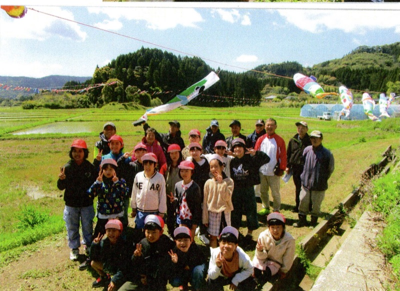 田園を泳ぐこいのぼりの前で児童たちと地域住民の記念撮影