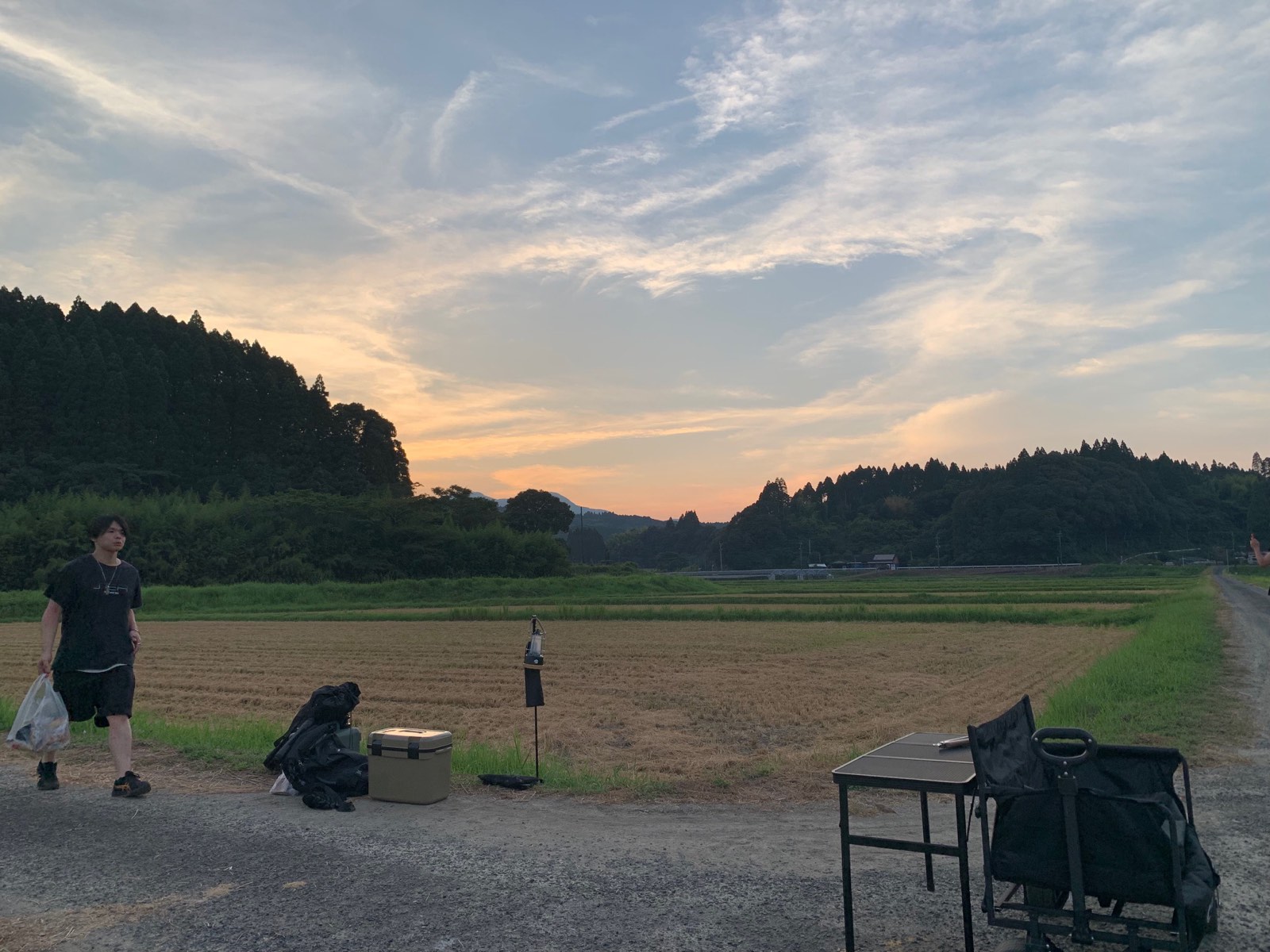 夕暮れの畦道に設営された焚き火の道具と、オレンジに染まる西の空